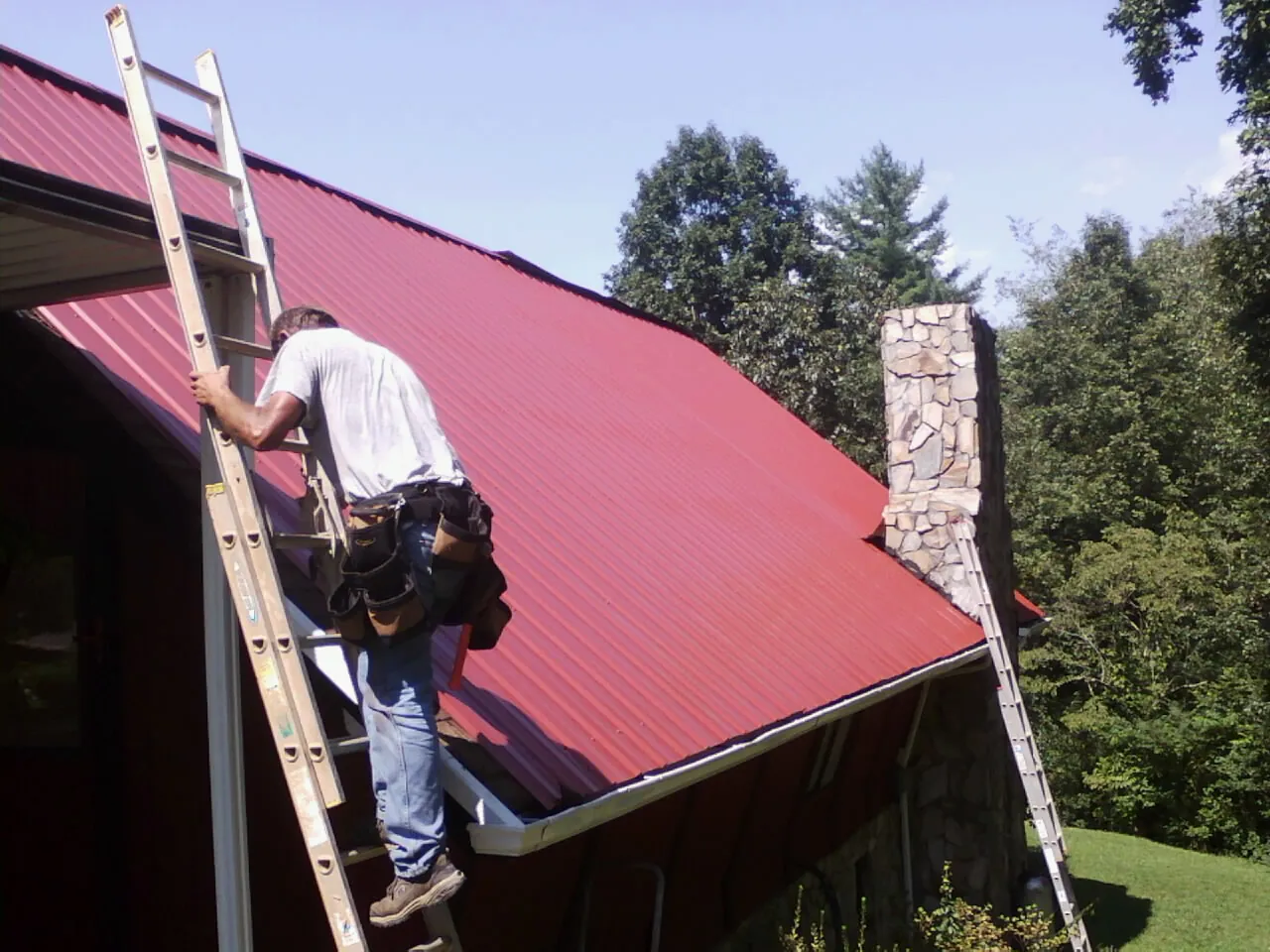 Roofer on a ladder beside a red metal roof and stone chimney — Soffit & Fascia Repair by Roofing Brothers in Edwards